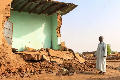 AFP An elderly man stands before the collapsed mudbrick wall of a house in the aftermath of flooding in the area of Messawi near Meroe in Sudan's Northern State on August 28, 2024.