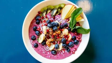 Getty Images A stock photo of a white bowl with purple acai paste and fruits including berries, banana, pomegranate seeds, kiwi, and mint leaves and nuts 