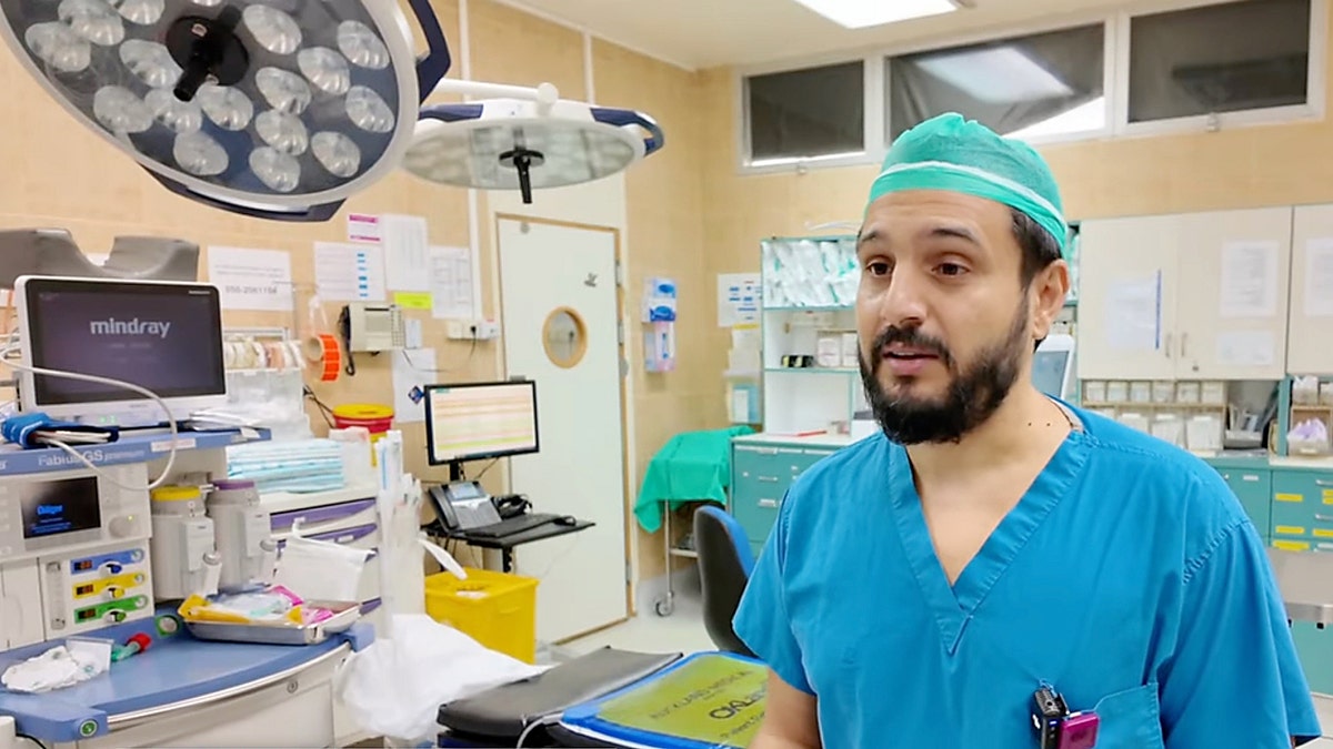Surgeon in blue scrubs speaks inside a brightly lit operating room with medical equipment behind him.
