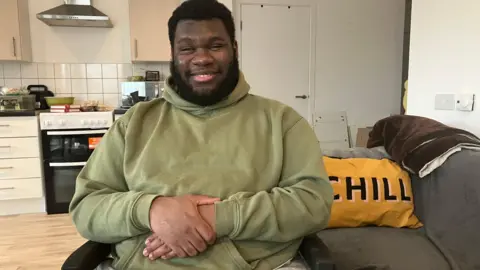 Young man with dark hair and a beard sits facing the camera, wearing a green hooded sweatshirt