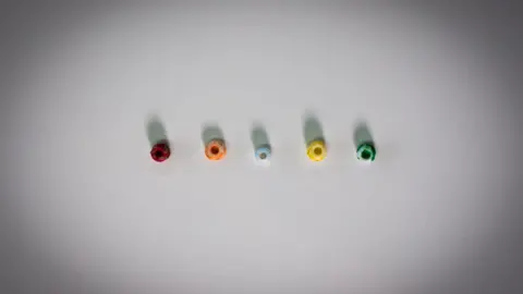 Kevin Church/BBC News Five small round beads arranged horizontally on a white surface.  Each bead has a hole in the centre.  They are multicoloured, from left to right, they are dark red,  orange, light blue, yellow and green. 